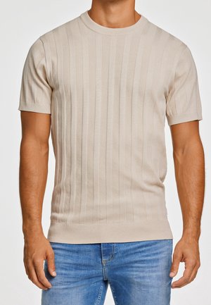 Mann trägt ein kurzärmeliges beiges Strickshirt mit vertikalen Streifen und blaue Jeans vor einfarbigem Hintergrund.