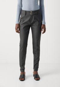 Zwarte op maat gemaakte broek met een gladde textuur, voorzien van twee decoratieve knopen bij de taille en taps toelopende enkels, gecombineerd met zwarte slipper sandalen.