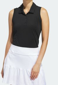 Polo noir sans manches avec col et quatre boutons, associé à une jupe blanche évasée ornée d'un volant texturé à l'ourlet.