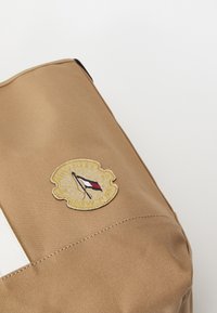 Brązowa torba tekstylna z wyraźnie naszytą łatą przedstawiającą jacht oraz napisem "Tommy Hilfiger New York", dopełniona białym paskiem.