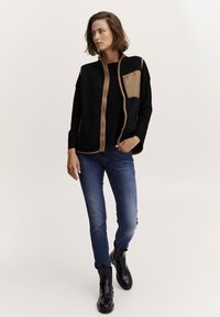 Gilet in pile nero con dettagli marroni, colletto alto, zip frontale e tasca a toppa. Abbinato a jeans blu e stivaletti neri.