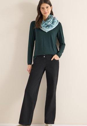 Tummanvihreä pitkähihaista neulepusero, yhdistetty mustiin leveälahkeisiin housuihin. Mallilla on vaaleansininen kuvioitu huivi kaulan ympärillä.