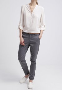 Blouse crème clair à boutons avec manches retroussées, associée à un pantalon gris slim et des chaussures blanches sans lacets, créant un look décontracté.