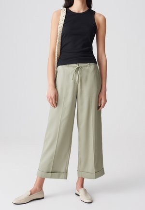 Kvinde iført en sort ærmeløs top, lysegrønne vidde bukser med snøre, cremefarvede loafers og en beige hæklet skuldertaske.