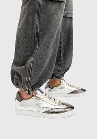 Metallic silver sneakers with a textured finish and contrasting dark accents, paired with gray elastic cuffed pants.