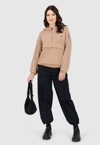 Felpa beige con zip e tasca frontale, abbinata a pantaloni cargo neri larghi e stivali neri grossi. Il modello tiene in mano una piccola borsa nera.