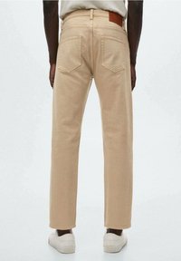 Pantalones chinos de algodón beige con diseño de pierna recta, que cuentan con dos bolsillos traseros, un parche de cuero marrón en la cinturilla y zapatillas blancas.