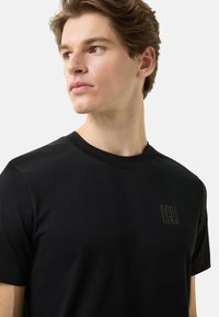 Černé přiléhavé tričko z bavlny s kulatým výstřihem, krátkými rukávy a nenápadným reliéfním logem na hrudi.