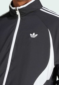 Svart sportjacka med hög krage, vitt blixtlås och kontrasterande vita detaljer. Har en broderad logga och slanka designdrag.