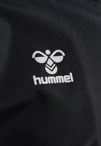 White hummel logo sa stiliziranim puhcem i tekstom na crnoj tkanini u pozadini.