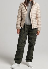 Béžová péřová bunda, šedý svetr a tmavě zelené cargo kalhoty s maskáčovým vzorem. Bílé sneakers, volný střih s kapsami na kalhotách. Neformální venkovní oblečení.