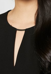 Svart klänning med V-formad halsringning, som har en liten metallisk DKNY-accent vid kragen. Mjuk textur, skräddarsydd passform.