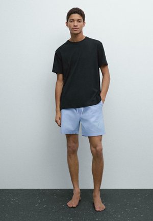 T-shirt en coton noir associé à un short bleu clair. Le short est doté d'une taille élastique avec cordon de serrage et d'une coupe décontractée. Modèle pieds nus.