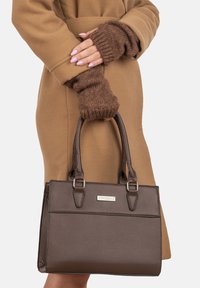 Bolso de mano de textura marrón con dos asas, forma rectangular, hardware plateado y placa de marca metálica. Lleva puesto un abrigo marrón claro.