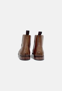 Botas de tobillo de cuero marrón con un acabado suave, detalles decorativos de brogue y una cinta roja en la parte posterior. Tacones de madera resistentes.