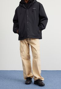 Person som bär en svart huva jacka, beige cargobyxor och svarta sneakers, som står på en blå matta med händerna i jackfickorna.