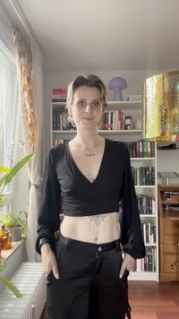 Schwarzes, kurzes Oberteil mit langen Ärmeln und V-Ausschnitt, kombiniert mit schwarzen Hosen. Sichtbare Haut im Bereich des Bauches, mit dekorativen Tattoos.