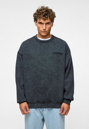 Donkergrijze crewneck sweatshirt van textuurstof, met dropped schouders en een subtiele logo op de borst. Gecombineerd met lichtblauwe jeans.