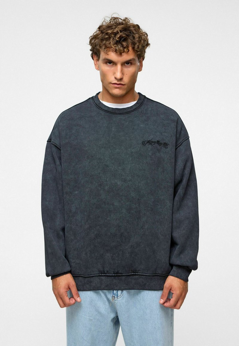 Donkergrijze crewneck sweatshirt van textuurstof, met dropped schouders en een subtiele logo op de borst. Gecombineerd met lichtblauwe jeans.