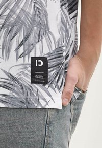 Zbliżenie ręki w kieszeni dżinsów, noszącej białą koszulę w czarny tropikalny wzór liści oraz czarną metkę marki dżinsowej.