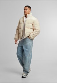 Kremfarget pufferjakke med en myk, teksturert overflate; høy krage; glidelås; kombinert med lys blå jeans og mønstrede sneakers.