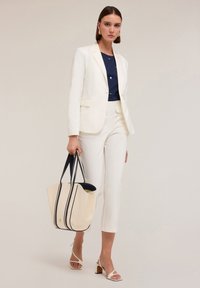 Blazer blanc ajusté sur un haut marine, associé à un pantalon cropped blanc. Tenant un sac fourre-tout beige avec des straps navy. Sandales à talons blocs.