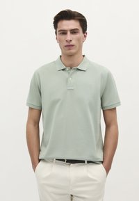 Ung mand iført en lys grøn kortærmet polo t-shirt og hvide bukser, stående med hænderne i lommerne mod en ensfarvet hvid baggrund.