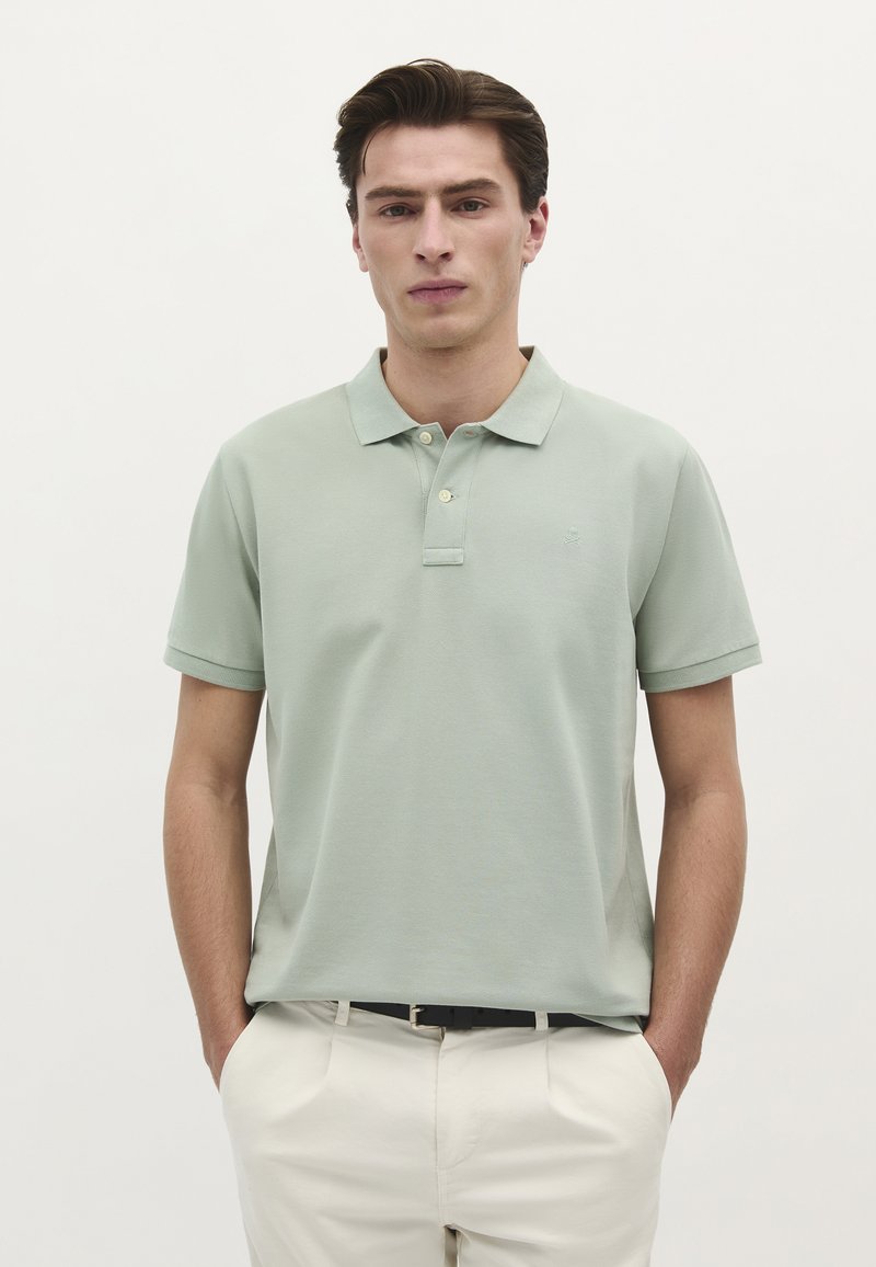 Ung mand iført en lys grøn kortærmet polo t-shirt og hvide bukser, stående med hænderne i lommerne mod en ensfarvet hvid baggrund.