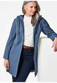 Lichtgewicht blauwe regenjas met een capuchon, gemaakt van gladde stof. Bevat een rits aan de voorkant en zijzakken, gedragen over een witte top en donkerblauwe jeans.