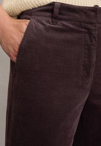 Bruine corduroy broek met verticale ribbelstructuur, voorzien van zakken en een relaxed fit, gedeeltelijk gezien op de taille met een hand in de zak.
