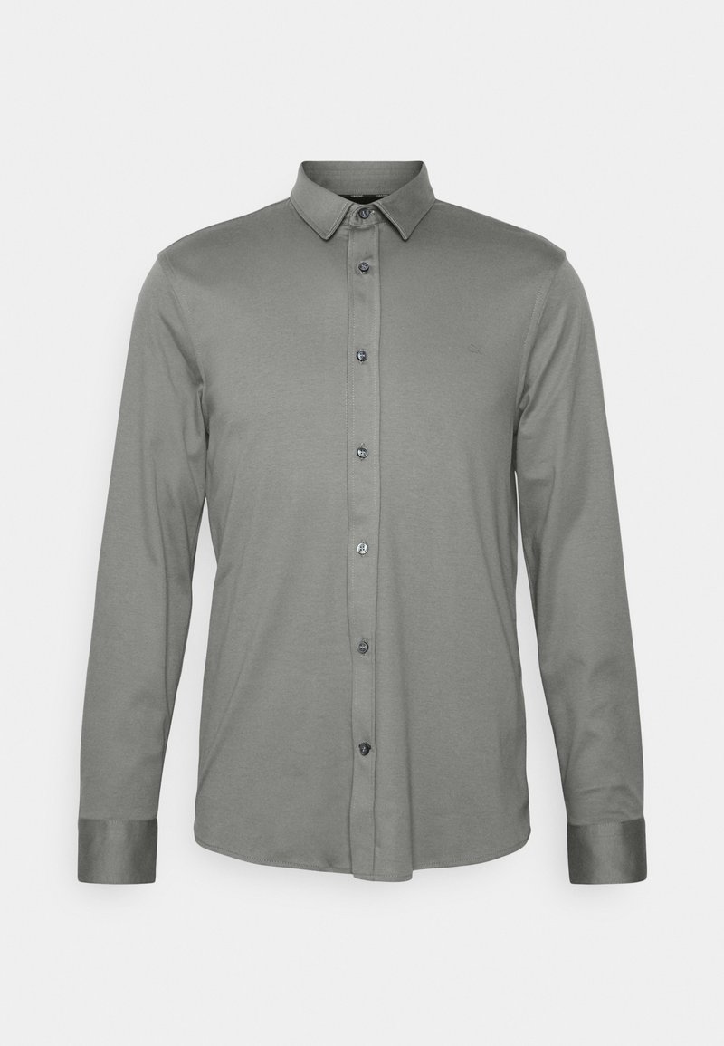 Chemise grise à manches longues avec boutons, col pointu et poignets boutonnés, présentée sur un fond blanc uni.