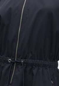 Μπλε εμπριμέ μπουφάν με κεντρικό ασημένιο φερμουάρ και ρυθμιζόμενο κορδόνι στη μέση, που δείχνει υφή υφάσματος και λεπτομέρειες ραφής.