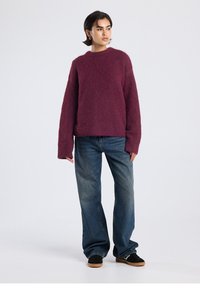 Burgundy knitted sweater with a crew neckline and oversized fit, paired with loose blue jeans and black shoes. Textured, warm appearance.