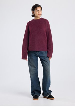 Maglione bordeaux lavorato a maglia con scollo rotondo e vestibilità oversize, abbinato a jeans blu larghi e scarpe nere. Aspetto strutturato e caldo.