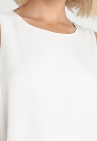 Femme portant une robe blanche texturée sans manches avec un col rond haut, laissant apparaître une partie de son cou et de ses épaules.