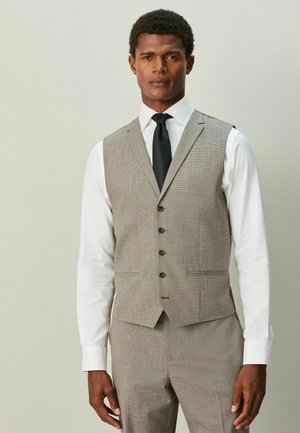 Mann trägt eine beige karierte Anzugweste und Hose über einem weißen Hemd mit schwarzer Krawatte, steht vor einem einfarbigen hellen Hintergrund.