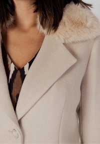 Abrigo de lana beige con un suave cuello de piel sintética, con solapa muesca, cierre de un solo botón y un acabado de tejido texturizado.