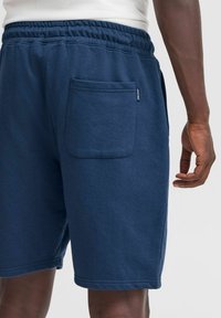 Marineblå bomulds shorts med elastisk talje, med en baglomme og glat tekstur, vist fra siden med en hånd hvilende på hoften.