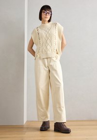 Suéter sin mangas de punto en color beige con patrones de trenzado texturizados, combinado con pantalones de pierna ancha en beige claro y botas de tobillo marrones con cordones.