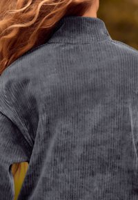 Haut en velours côtelé gris avec des côtes verticales, un col haut et une coupe décontractée. Les caractéristiques incluent une texture lisse et des manches amples.