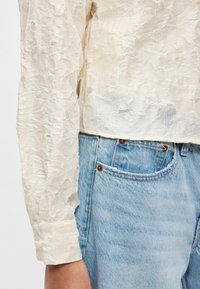 Lekka, fakturowana kremowa bluzka z marszczonym materiałem i długimi rękawami, zestawiona z jasnoniebieskimi dżinsami z kieszenią i metalowymi nity.