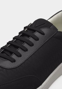 Μαύρο αθλητικό παπούτσι με δερμάτινη επένδυση υφής, μαύρα κορδόνια, ανάγλυφο λογότυπο στη γλώσσα και λευκή σόλα, εμφανίζεται σε κοντινή όψη.