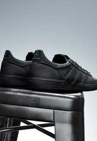 Baskets Adidas noires avec le logo Mercedes AMG au talon, placées sur un tabouret en métal noir devant un fond gris clair uni.