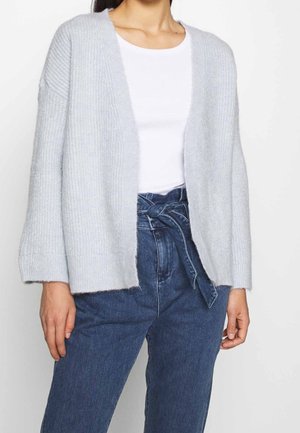 Vrouw draagt een lichtgrijze openvallende cardigan, een wit nauwsluitend topje en een hooggetailleerde blauwe paperbag jeans met een gebonden riem.