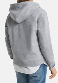 Grijze hoodie met een ontspannen pasvorm, zachte textuur en geribbelde mouwen. Draagbaar over een wit overhemd, met een lichte verlaging aan de zoom.