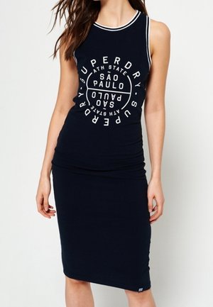 Kobieta w czarnej sukience bez rękawów z białym wykończeniem i logo São Paulo Superdry nadrukowanym na piersi.
