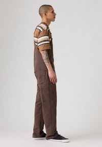 Man met kort haar en een getatoeëerde arm, gekleed in een bruine overall, een gestreept bruin en beige shirt en zwarte sneakers, staat zijwaarts tegen een grijze achtergrond.