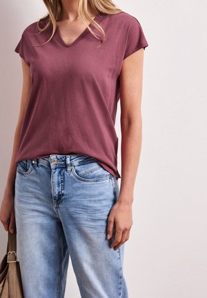 Femme portant un t-shirt mauve à manches courtes et col en V, légèrement rentré dans un jean en denim bleu clair, tenant un sac à main en cuir beige.