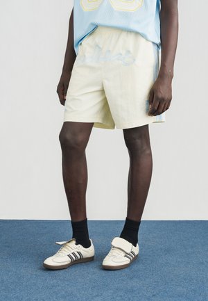 Lysegule shorts med blå accenter og broderet "Adidas"-logo, kombineret med cremefarvede sneakers med sorte striber og en struktureret rem.