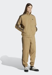 Beige tracksuit with black side stripes, zip-up jacket, elastic waistband, and cuffed pants. Black and white sneakers complete the outfit.